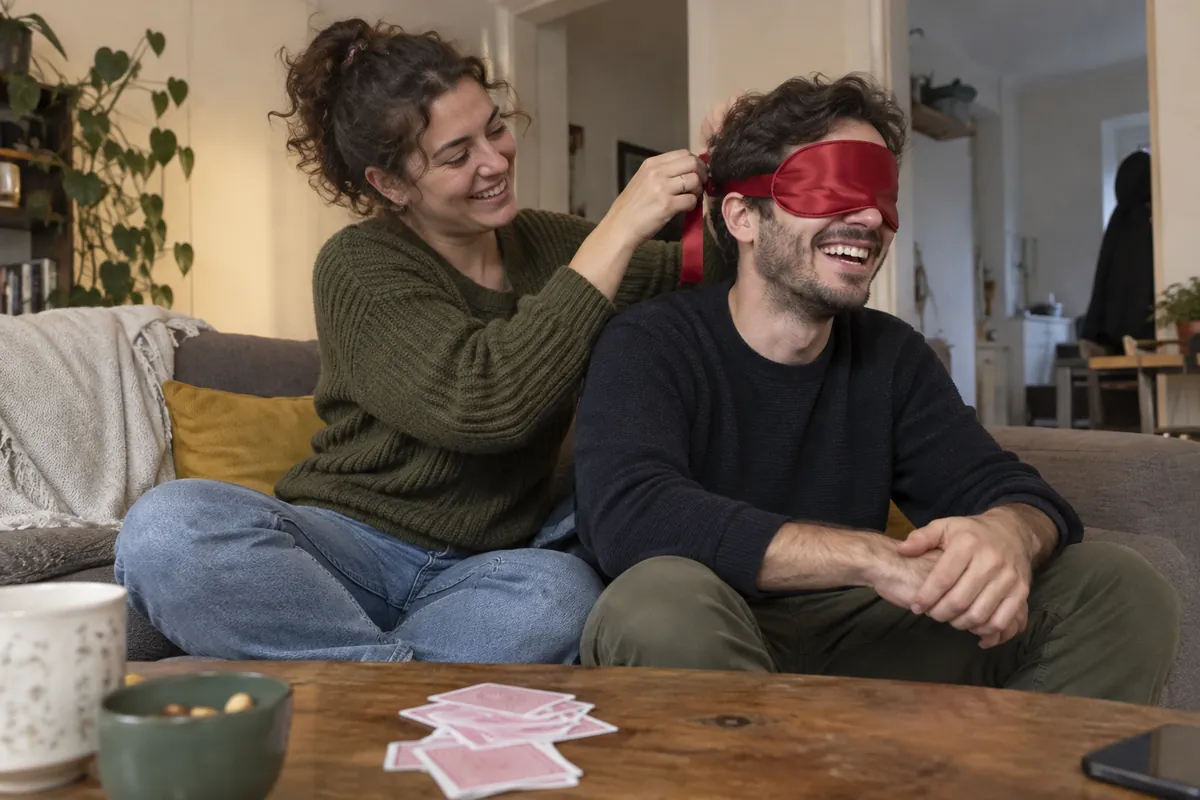 Woman ties a red satin blindfold over her laughing partner's eyes on a sofa, playing cards on the coffee table