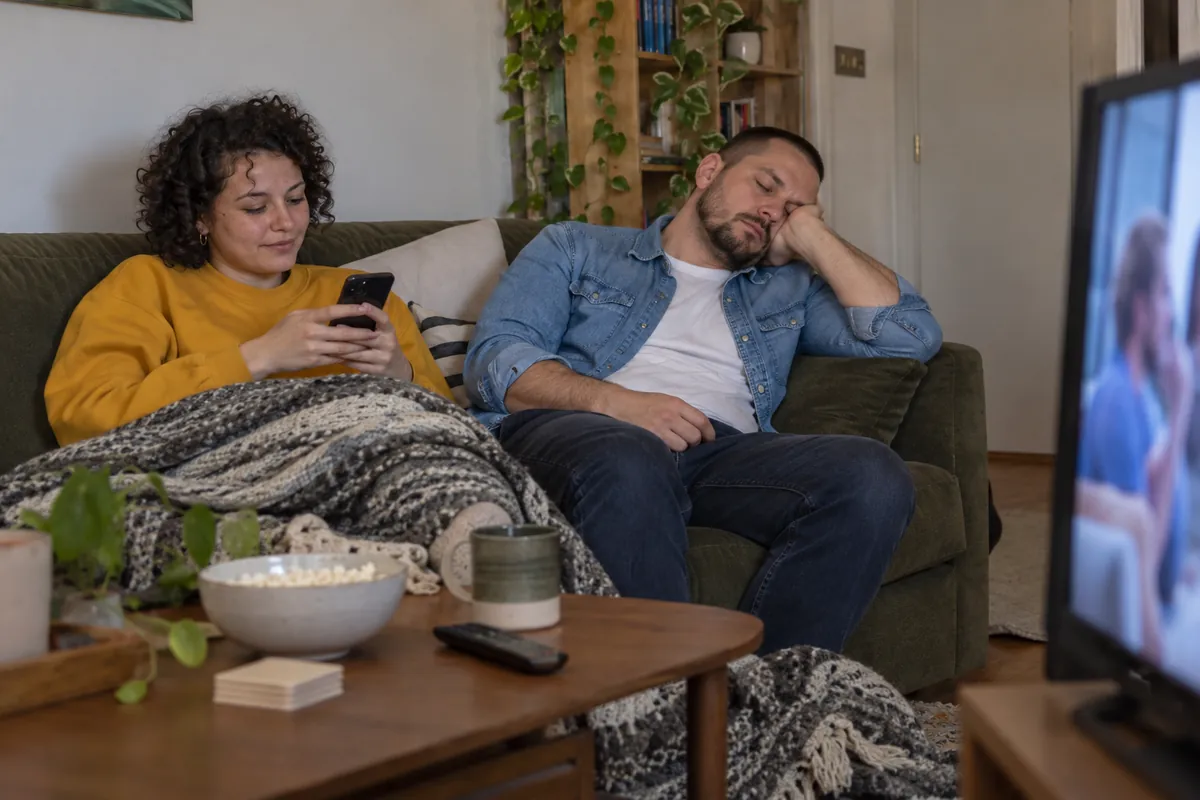 He has dozed off on the sofa while she scrolls her phone under a blanket, the TV playing to nobody