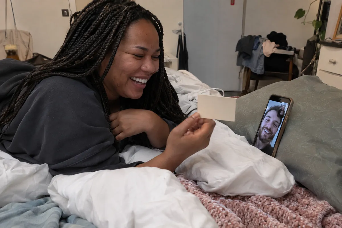 Woman with long braids lies on her bed laughing on a video call, holding a card up to her boyfriend on the propped-up phone