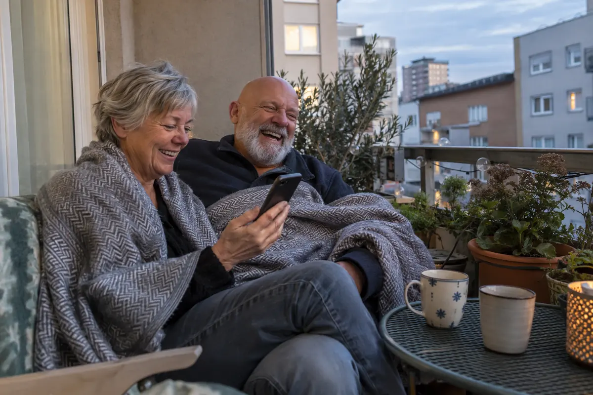 Older couple under a shared blanket on a balcony laughing at a question on his phone, mugs on a small table