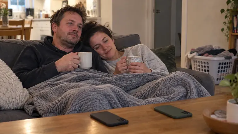 Couple under one blanket on a sofa holding mugs, both phones lying face down on the coffee table