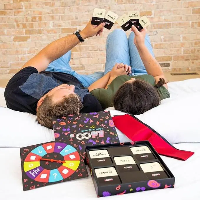 Couple lying on their backs on a bed holding Loopy cards up, the box, spinner board and red blindfold beside them