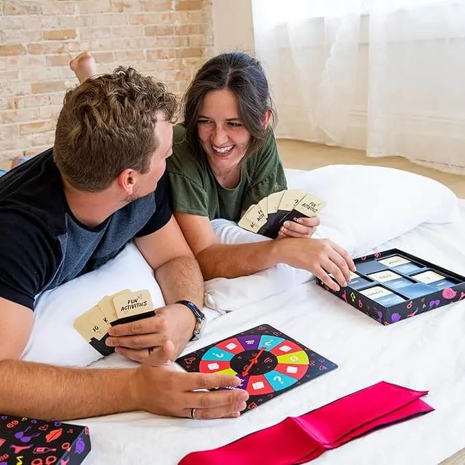 Couple lying on a bed laughing over Loopy cards, with the spinner board, card tray and a pink blindfold in front