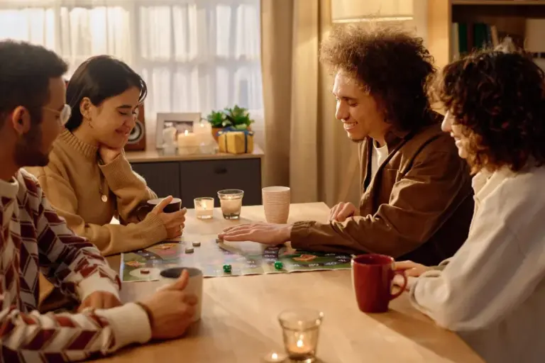 Four friends around a wooden table playing a board game, with mugs, glasses and a lit candle